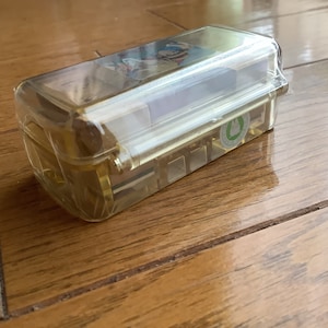 May include: A rectangular, transparent plastic container with a hinged lid. The container is filled with small, rectangular objects. A sticker with a green emblem is visible on the side. The container is sitting on a wooden surface.