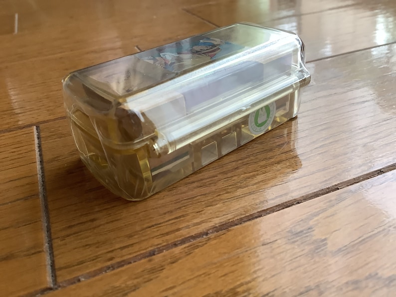 May include: A rectangular, transparent plastic container with a hinged lid. The container is filled with small, rectangular objects. A sticker with a green emblem is visible on the side. The container is sitting on a wooden surface.