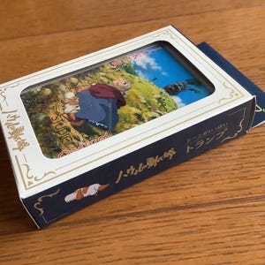 Pu&ograve; includere: Un set di carte da gioco in scatola con un'illustrazione colorata di un film d'animazione. La scatola &egrave; bianca con finiture dorate e testo giapponese. Un libretto blu &egrave; appoggiato accanto alla scatola su una superficie di legno.