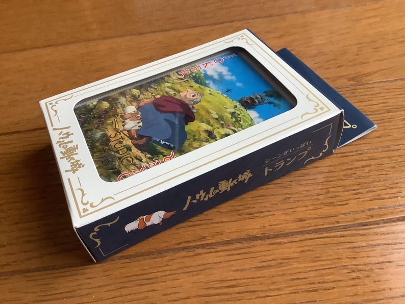 Pu&ograve; includere: Un set di carte da gioco in scatola con un'illustrazione colorata di un film d'animazione. La scatola &egrave; bianca con finiture dorate e testo giapponese. Un libretto blu &egrave; appoggiato accanto alla scatola su una superficie di legno.