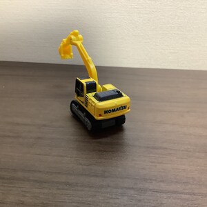 May include: A yellow Komatsu toy excavator with a black top and tracks. The excavator arm is raised. The toy is on a brown surface.