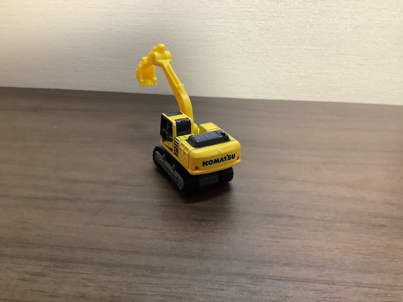 May include: A yellow Komatsu toy excavator with a black top and tracks. The excavator arm is raised. The toy is on a brown surface.