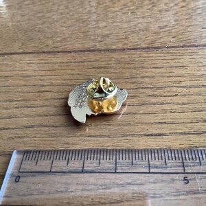 May include: A gold-toned metal lapel pin shown from the back, featuring a butterfly clutch fastener. The pin is positioned next to a clear plastic ruler that shows a measurement of approximately 1.5 inches in width for the pin's base.