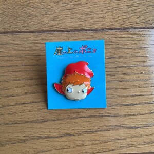May include: A metal enamel pin featuring the character Ponyo with orange hair and a red hood, attached to a square blue backing card. The card displays the Japanese text 崖の上のポニョ and the English subtitle Ponyo on the Cliff by the Sea.
