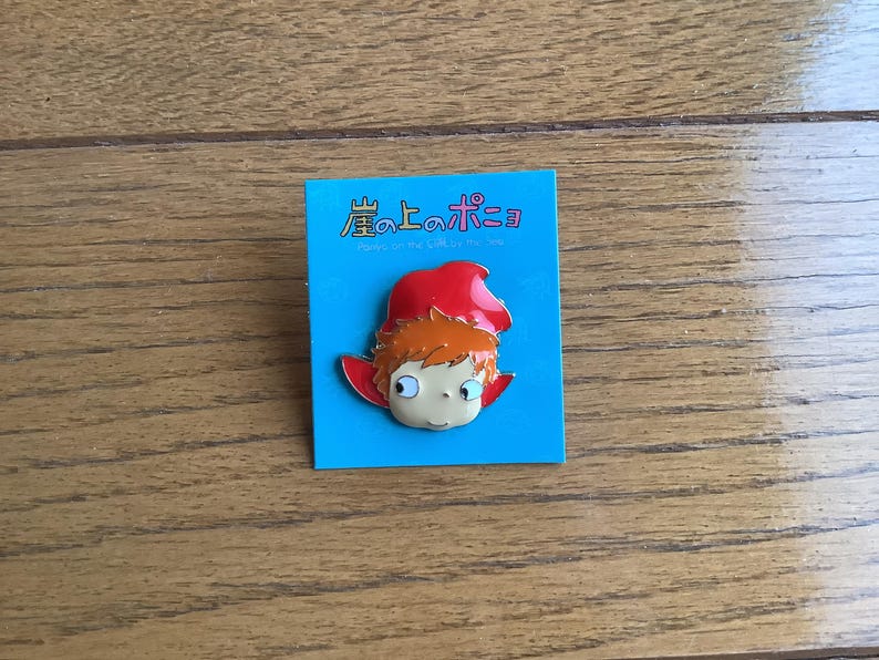 May include: A metal enamel pin featuring the character Ponyo with orange hair and a red hood, attached to a square blue backing card. The card displays the Japanese text 崖の上のポニョ and the English subtitle Ponyo on the Cliff by the Sea.