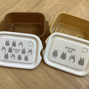 May include: Two rectangular lunch boxes with lids featuring the text "MY NEIGHBOR TOTORO" and Totoro character illustrations. The boxes are brown and tan, with white lids. The lids have a button closure.