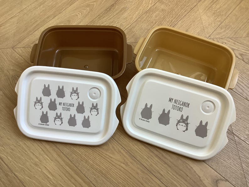 May include: Two rectangular lunch boxes with lids featuring the text "MY NEIGHBOR TOTORO" and Totoro character illustrations. The boxes are brown and tan, with white lids. The lids have a button closure.