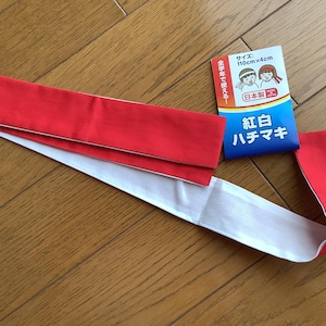 May include: A long, rectangular red and white fabric headband, known as a hachimaki, accompanied by a product label featuring illustrations of two people wearing the headband and text indicating dimensions of 110cm x 4cm and Japanese origin.
