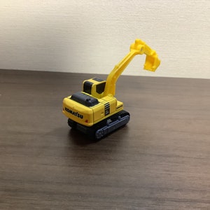 May include: A yellow and black Komatsu toy excavator with a raised arm and bucket. The toy has black treads and the Komatsu logo on the back. The excavator is on a brown surface.