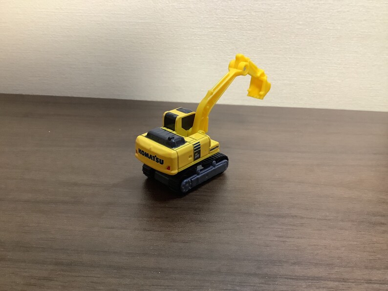 May include: A yellow and black Komatsu toy excavator with a raised arm and bucket. The toy has black treads and the Komatsu logo on the back. The excavator is on a brown surface.