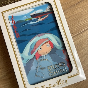 May include: A playing card featuring a scene from Ponyo on the Cliff by the Sea, showing a character with pink hair under a light blue blanket, a ship on the ocean, the word JOKER in red, and the gold embossed text STUDIO GHIBLI in a white box.
