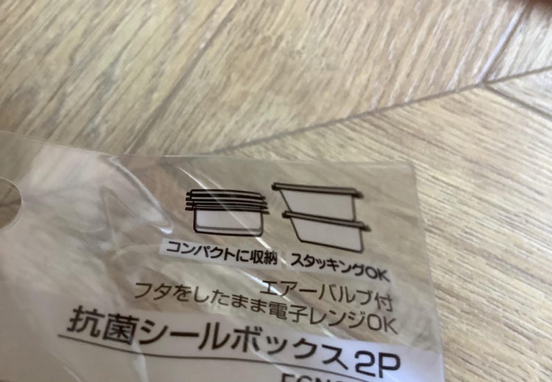 May include: A clear plastic package with Japanese text and illustrations of food storage containers. The text includes phrases like "compact storage" and "microwave safe." The image shows a close-up of the packaging.