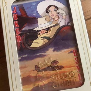 May include: A Studio Ghibli themed playing card set featuring a Joker card with an illustration of a woman and a pig in a plane. The card set is in a white and gold box with the words "Joker" and "Studio Ghibli".