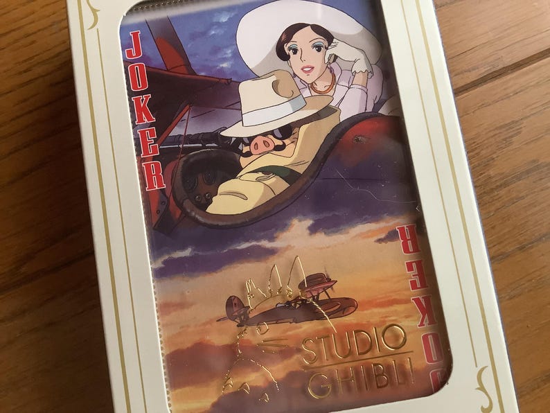 May include: A Studio Ghibli themed playing card set featuring a Joker card with an illustration of a woman and a pig in a plane. The card set is in a white and gold box with the words "Joker" and "Studio Ghibli".