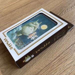 May include: A deck of playing cards featuring a whimsical illustration of Totoro from the animated film My Neighbor Totoro. The card box is white and brown with Japanese text and a Totoro graphic.