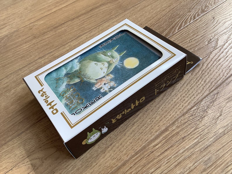 May include: A deck of playing cards featuring a whimsical illustration of Totoro from the animated film My Neighbor Totoro. The card box is white and brown with Japanese text and a Totoro graphic.