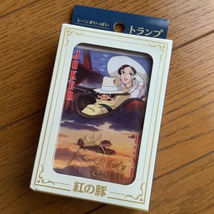 May include: A deck of playing cards featuring a scene from the animated film "Porco Rosso." The card box is white with a clear window showing the card design. The design includes a woman in a hat and a pig piloting a plane.