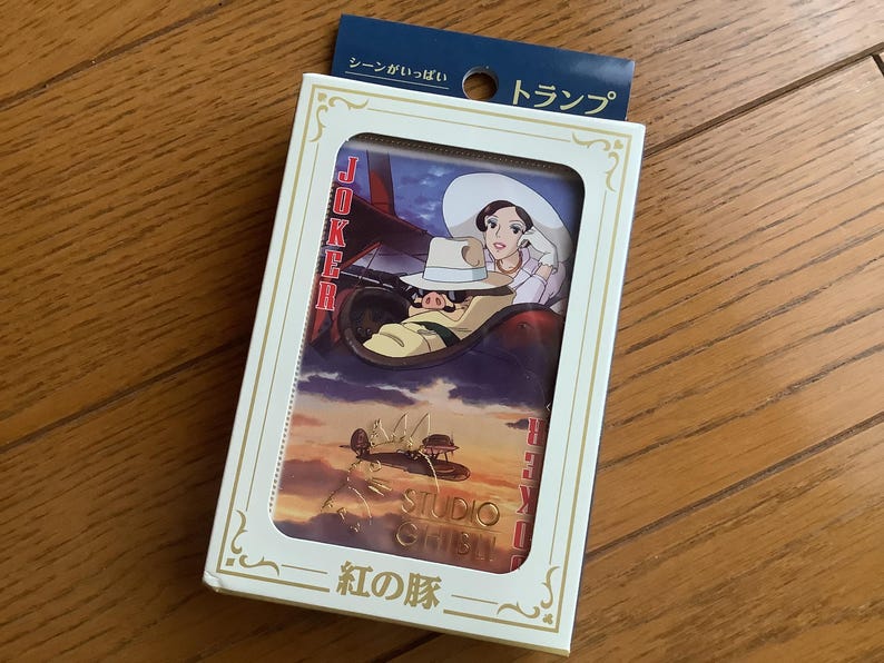 May include: A deck of playing cards featuring a scene from the animated film "Porco Rosso." The card box is white with a clear window showing the card design. The design includes a woman in a hat and a pig piloting a plane.
