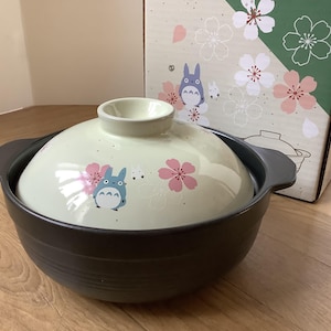 May include: A black and cream-colored ceramic pot with a lid featuring a cartoon character and pink floral designs. The pot has two handles and sits in front of a box with similar illustrations.