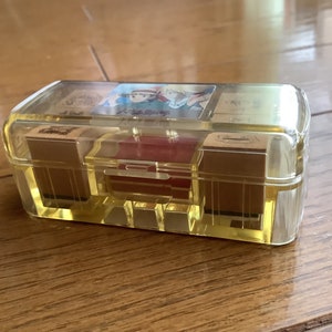 May include: A clear, rectangular box with a yellow tint, containing several small, rectangular stamps. The box has a hinged lid and a red interior compartment. The stamps appear to have illustrations on them. The box is sitting on a wooden surface.
