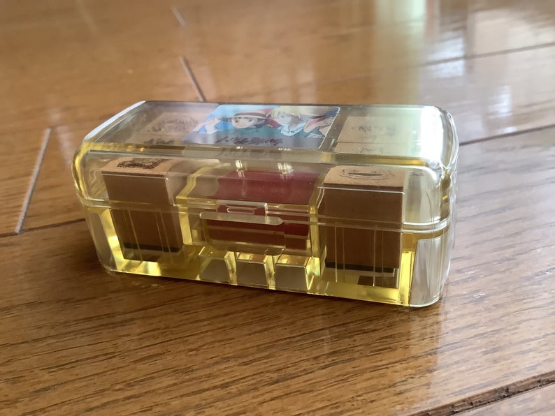 May include: A clear, rectangular box with a yellow tint, containing several small, rectangular stamps. The box has a hinged lid and a red interior compartment. The stamps appear to have illustrations on them. The box is sitting on a wooden surface.