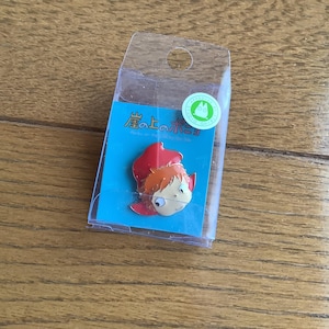 May include: A metal enamel pin featuring a cartoon character with orange hair and a red hood, mounted on a blue backing card inside a clear plastic display box. The card displays Japanese text and a small green circular sticker with a white character icon.