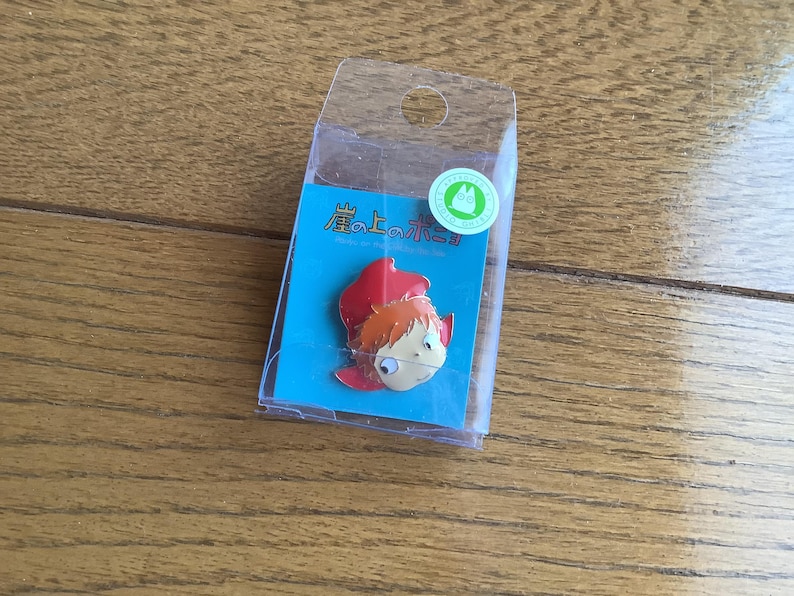 May include: A metal enamel pin featuring a cartoon character with orange hair and a red hood, mounted on a blue backing card inside a clear plastic display box. The card displays Japanese text and a small green circular sticker with a white character icon.