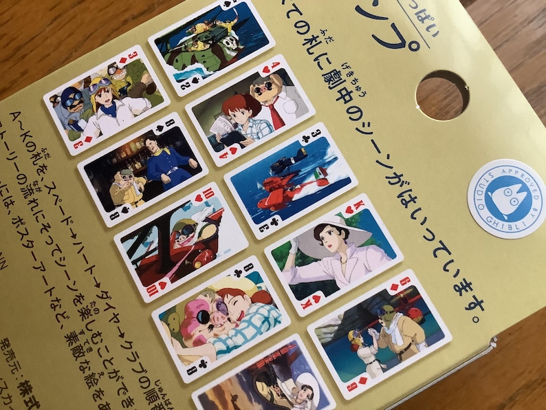 May include: Playing cards featuring illustrations from a Japanese animated film. The cards display various scenes and characters, with the Studio Ghibli seal visible. The cards are arranged on a gold-colored surface.