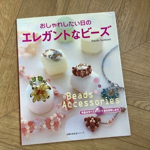 May include: A softcover craft book titled "おしゃれしたい日のエレガントなビーズ" and "Beads Accessories" by Paolo Bottoni. The cover features photographs of various beaded rings, necklaces, and bracelets displayed on white cylindrical stands.