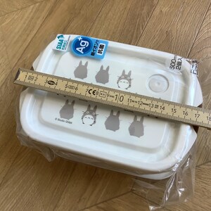 May include: White rectangular food container with a lid featuring gray Totoro character illustrations. The container is approximately 7 inches long. The packaging includes Japanese text and a blue and white label.