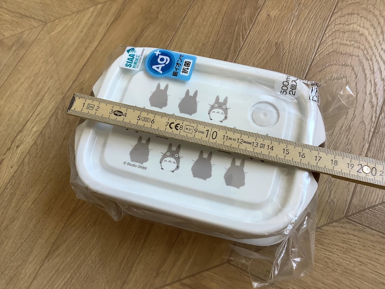 May include: White rectangular food container with a lid featuring gray Totoro character illustrations. The container is approximately 7 inches long. The packaging includes Japanese text and a blue and white label.