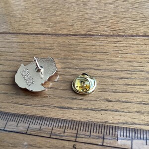 May include: A gold-toned metal lapel pin back featuring an embossed logo and a separate circular clutch fastener, positioned next to a ruler showing measurements in inches to provide a clear sense of the scale of these small metal components.