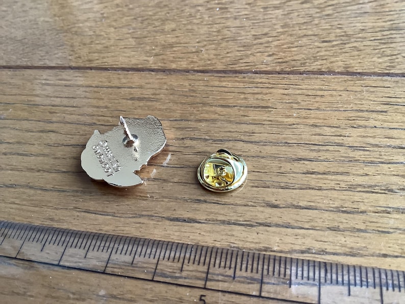 May include: A gold-toned metal lapel pin back featuring an embossed logo and a separate circular clutch fastener, positioned next to a ruler showing measurements in inches to provide a clear sense of the scale of these small metal components.