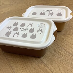 May include: Two brown and white rectangular food containers with lids. The lids are white and feature a repeating pattern of gray Totoro characters and the text "MY NEIGHBOR TOTORO". The containers are on a wooden surface.