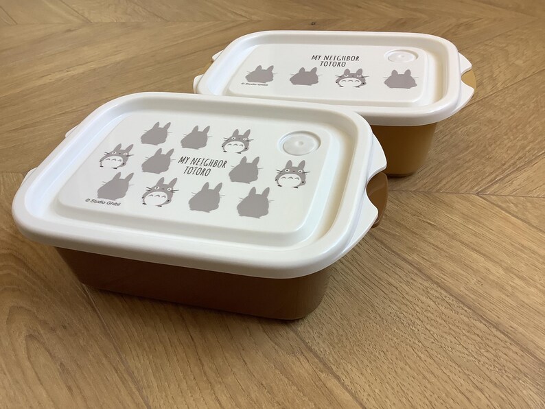 May include: Two brown and white rectangular food containers with lids. The lids are white and feature a repeating pattern of gray Totoro characters and the text "MY NEIGHBOR TOTORO". The containers are on a wooden surface.