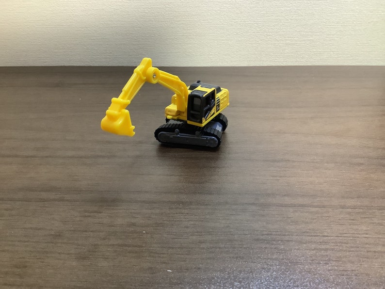 May include: A yellow and black toy excavator with a movable arm and bucket. The toy is on a wooden surface. The excavator has black tracks and a detailed cabin. A fun toy for kids.