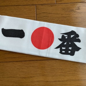 May include: White headband featuring a red circle and black Japanese characters. The headband is made of soft, white fabric. The Japanese characters and the red circle are printed on the front of the headband.