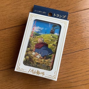 Pu&ograve; includere: Un mazzo di carte da gioco in una scatola bianca. La carta visibile mostra una scena con un personaggio in abito blu e scialle rosso, con un castello sullo sfondo. La scatola ha testo giapponese.