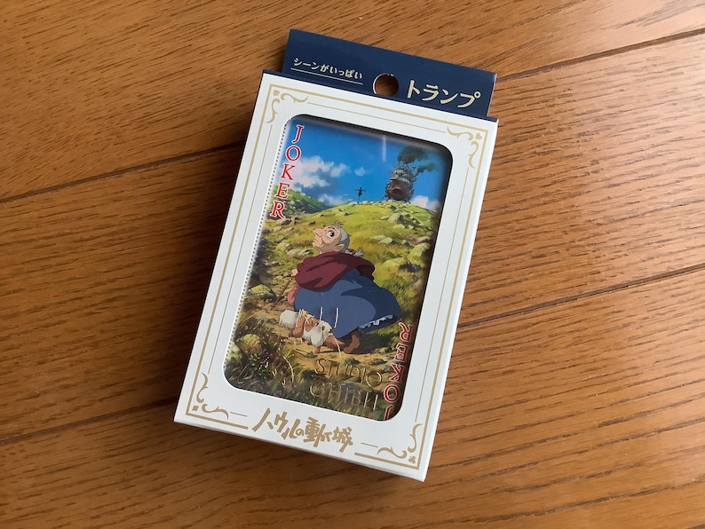 Pu&ograve; includere: Un mazzo di carte da gioco in una scatola bianca. La carta visibile mostra una scena con un personaggio in abito blu e scialle rosso, con un castello sullo sfondo. La scatola ha testo giapponese.
