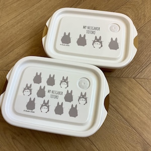 May include: Two white rectangular lunch boxes with rounded corners. Each lid features a gray illustration of the character Totoro from My Neighbor Totoro, along with the text "MY NEIGHBOR TOTORO". The boxes have brown accents.