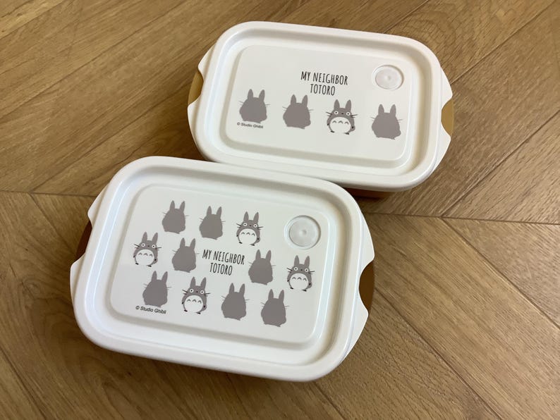 May include: Two white rectangular lunch boxes with rounded corners. Each lid features a gray illustration of the character Totoro from My Neighbor Totoro, along with the text "MY NEIGHBOR TOTORO". The boxes have brown accents.