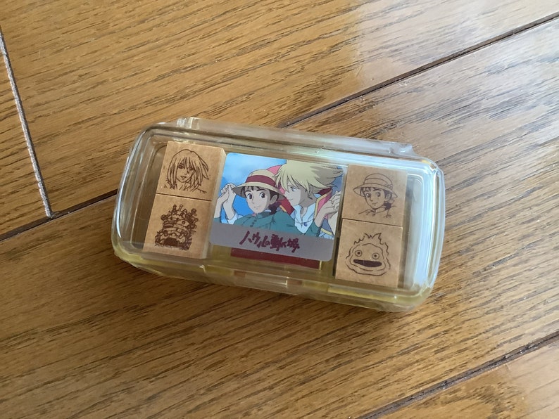 May include: A clear plastic case containing wooden stamps with illustrations of characters from the animated film "Howl's Moving Castle." The stamps are arranged inside the case, with a colorful image of the film's characters in the center.