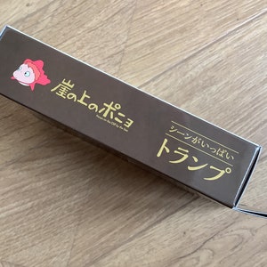 May include: A rectangular brown cardboard box featuring a small pink cartoon fish character on the left. The text reads "崖の上のポニョ", "Ponyo on the Cliff by the Sea", and "シーンがいっぱい トランプ" in gold lettering. The box has a flap extending from the right side.