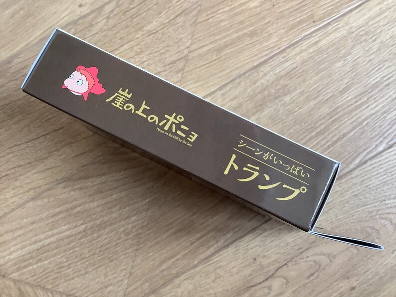 May include: A rectangular brown cardboard box featuring a small pink cartoon fish character on the left. The text reads "崖の上のポニョ", "Ponyo on the Cliff by the Sea", and "シーンがいっぱい トランプ" in gold lettering. The box has a flap extending from the right side.
