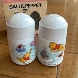 Op de afbeelding: Een paar witte cilindrische zout- en peperstrooiers met Winnie de Poeh-figuren, Knorretje en een kasteelafbeelding op een lichtblauwe achtergrond. De strooiers hebben witte koepelvormige deksels en staan voor een doos met de tekst SALT & PEPPER SET.