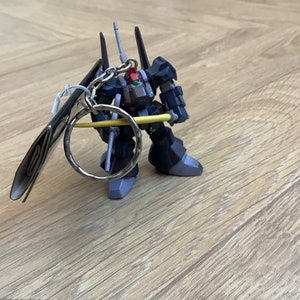 May include: A dark gray robot keychain with a silver key ring and a yellow bar. The robot has black and gray accents, with a red detail. The keychain is on a light brown wooden surface.