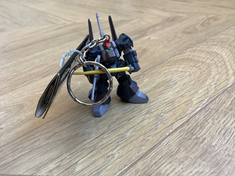 May include: A dark gray robot keychain with a silver key ring and a yellow bar. The robot has black and gray accents, with a red detail. The keychain is on a light brown wooden surface.