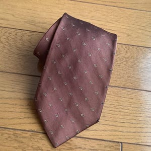 May include: A brown necktie featuring a subtle diagonal herringbone weave pattern and a repeating motif of small, light-colored, curved shapes arranged in orderly rows across the surface of the fabric, creating a textured and patterned appearance.