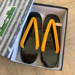May include: A pair of black geta sandals with bright yellow straps, presented in a striped box. The sandals have a glossy finish and are designed for traditional Japanese wear.
