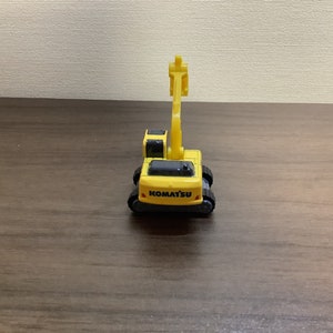 May include: A yellow Komatsu toy excavator with black tracks and a movable arm. The toy has the Komatsu logo in black on the front. The toy is on a brown wooden surface.